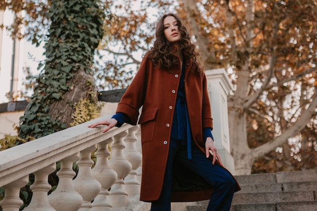 Foto grátis linda elegante e sorridente mulher magra com cabelo encaracolado andando nas escadas da rua, vestida com um casaco marrom quente e terno azul, outono na moda estilo de rua
