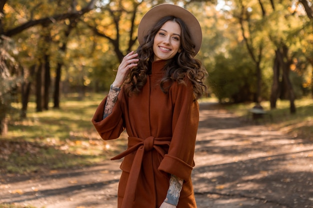 Linda elegante e sorridente mulher magra com cabelo encaracolado caminhando no parque, vestida com um casaco marrom quente, estilo de rua da moda outono
