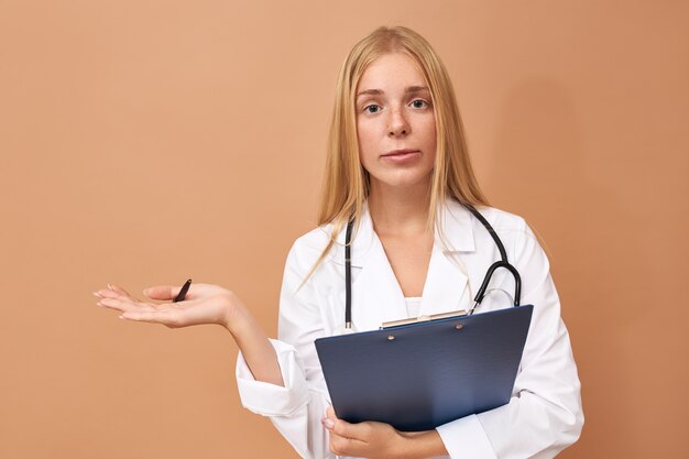 Linda elegante jovem cirurgiã em vestido branco segurando caneta e prancheta gesticulando, contando prós e contras da operação para seu paciente, discutindo riscos e efeitos colaterais