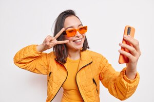 Linda elegante mulher asiática leva selfie no celular faz o sinal v sorrisos tem rosto positivo usa óculos de sol laranja e jaqueta isolada sobre uma parede branca. estilo de vida moderno