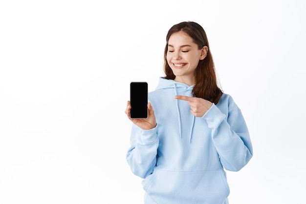 Linda estudante adolescente apontando e mostrando o aplicativo móvel de tela vazia do smartphone sorrindo satisfeito em pé contra o fundo branco