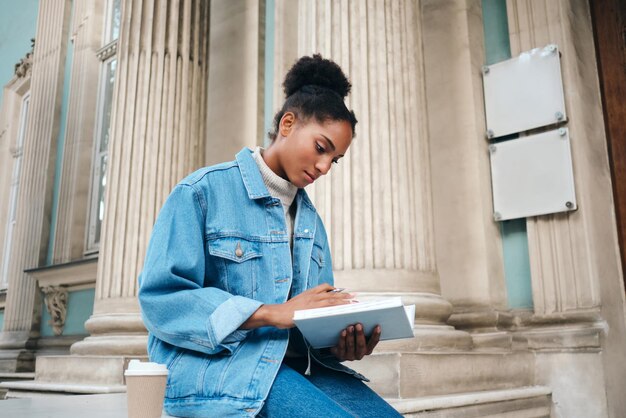 Linda estudante afro-americana casual em jaqueta jeans com livro estudando cuidadosamente ao ar livre