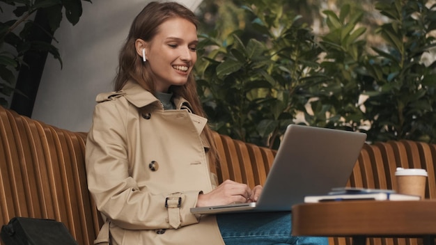 Foto grátis linda estudante em fones de ouvido sem fio parecendo feliz tendo palestra por videochamada no laptop estudando on-line no café ao ar livre