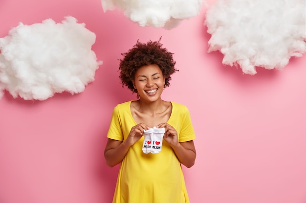 Linda feliz mulher grávida étnica com grande abdômen segurando botinhas se preparando para a maternidade vestida de vestido amarelo