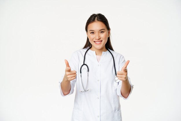 Linda fêmea médica enfermeira asiática em uniforme mostrando os polegares e sorrindo satisfeito, assegure o paciente ...