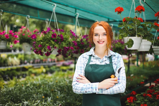 Linda florista sorridente em pé de avental com uma tesoura de jardim na mão. moça alegremente olhando na câmera enquanto trabalha com flores