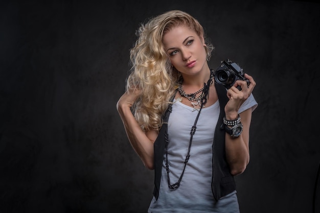 Linda fotógrafa loira encaracolada vestida com uma camiseta branca e colete usa muitos acessórios e relógio de pulso, posando com uma câmera em um estúdio. isolado em plano de fundo texturizado escuro.