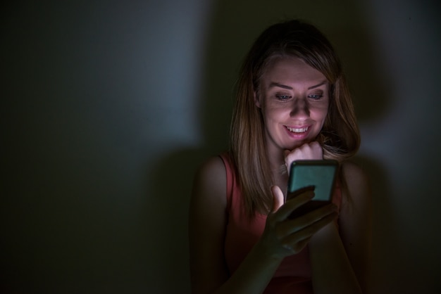 Linda garota à noite em um quarto escuro com telefone, mensagens de texto. Retrato de jovem mulher olhando a tela do telefone celular à noite