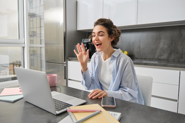 Foto grátis linda garota acena com as mãos para a câmera do laptop e diz olá aos colegas de trabalho, conecta-se à reunião de trabalho on-line
