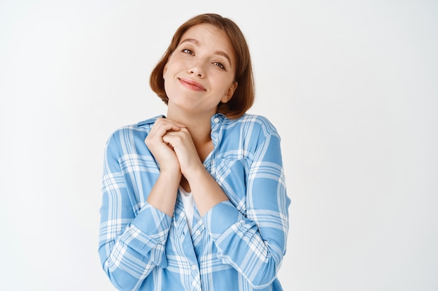 Linda garota admira algo fofo, suspirando e apertando as mãos, sorrindo e parecendo fascinada por algo lindo, parede branca