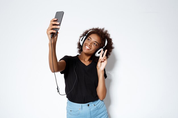 Linda garota africana em fones de ouvido, fazendo selfie sobre parede branca.