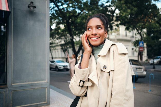 Linda garota afro-americana animada em um elegante casaco de trincheira falando alegremente no celular na rua da cidade