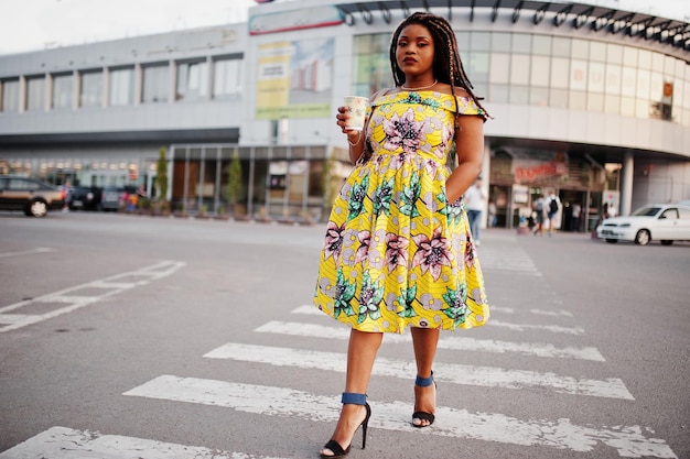 Foto grátis linda garota afro-americana de pequena altura com dreadlocks vestindo vestido amarelo colorido andando na faixa de pedestres contra o centro de comércio com uma xícara de café à mão