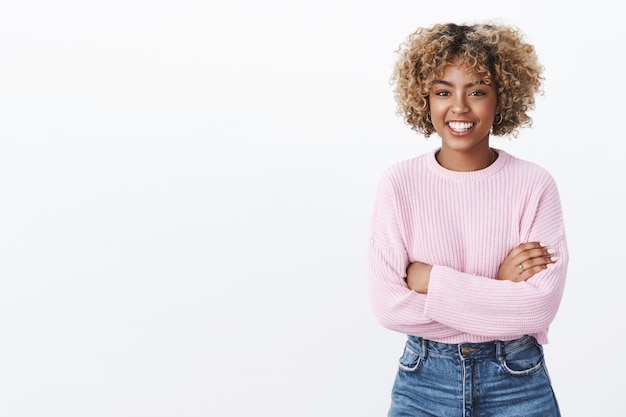 Foto grátis linda garota afro-americana entretida com uma conversa engraçada em pé, relaxada e despreocupada com as mãos cruzadas sobre o corpo, sorrindo amigavelmente para a câmera enquanto fala, vestindo um suéter roxo quente e moderno