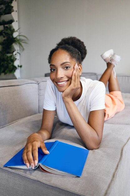 Linda garota afro-americana sorridente deitada no sofá com livro olhando alegremente na câmera em casa moderna