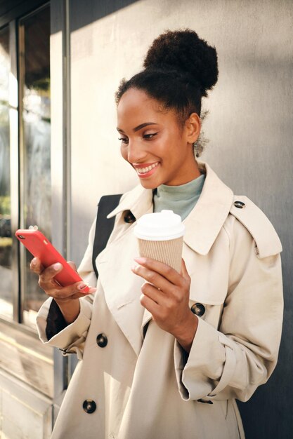 Linda garota afro-americana sorridente em um elegante casaco de trincheira com café para ir feliz usando o celular ao ar livre