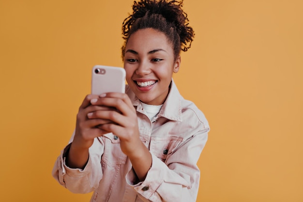 Linda garota afro-americana tirando foto com smartphone Vista frontal de uma mulher negra deslumbrante usando dispositivo digital em fundo laranja
