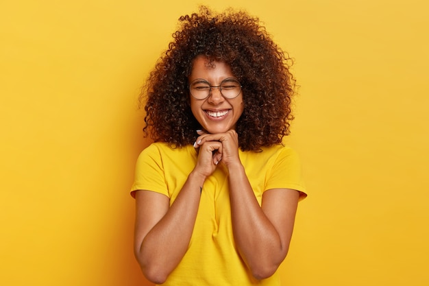 Foto grátis linda garota afro charmosa com cabelos cacheados, alegra a vida, mantém as mãos sob o queixo, sente-se radiante e satisfeita, tem aparência natural, veste uma camiseta amarela brilhante, posa em ambientes internos. conceito de felicidade