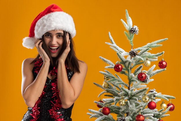 Linda garota alegre com chapéu de Natal com guirlanda no pescoço em pé perto da árvore de Natal isolada em fundo laranja