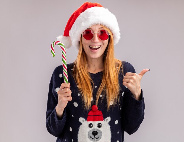 Linda garota alegre com chapéu de natal e óculos segurando um doce de natal, mostrando o polegar isolado no fundo branco