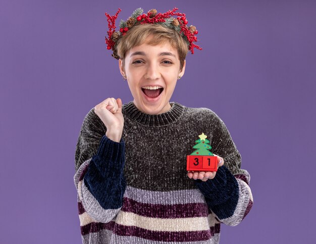 Linda garota alegre com coroa de flores na cabeça, segurando o brinquedo da árvore de Natal com data, olhando para a câmera, fazendo gesto de sim isolado no fundo roxo