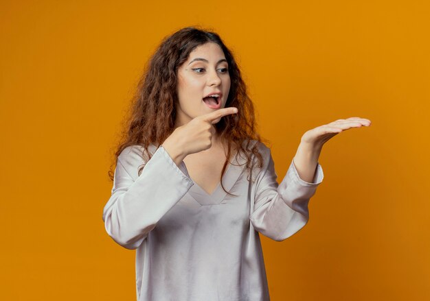 Linda garota alegre fingindo e apontando para algo isolado na parede amarela