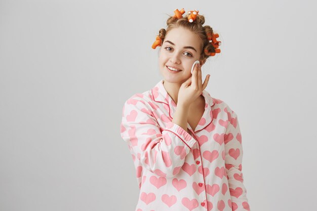 Linda garota aplique limpador antes de dormir com uma almofada de algodão, usando babadores de cabelo e pijama