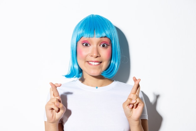 Linda garota asiática animada de peruca azul mordendo o lábio e olhando esperançosa para a câmera