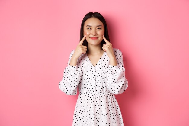 Linda garota asiática corada cutucando as bochechas, sorrindo feliz para a câmera, usando um vestido branco romântico, em pé sobre um fundo rosa