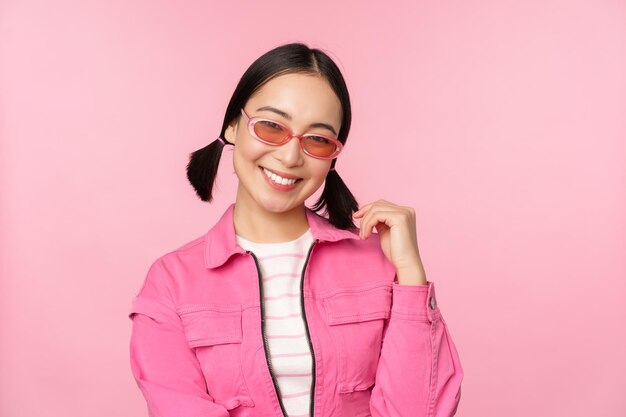 Linda garota asiática em sunlgasses sorrindo para a câmera posando contra o fundo rosa do estúdio