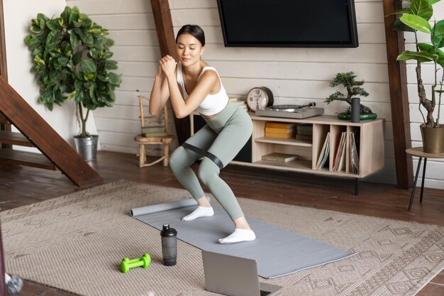 Linda garota asiática fitness em casa fazendo agachamento de treino com alongamento de corda elástica nas pernas em pé.
