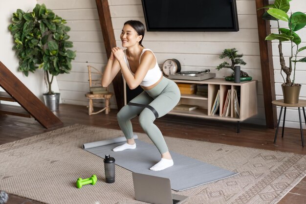 Linda garota asiática fitness em casa fazendo agachamento de treino com alongamento de corda elástica nas pernas em pé.