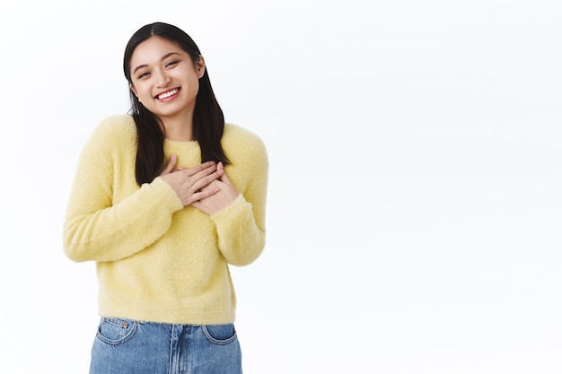 Linda garota asiática linda em um suéter amarelo, aperta as mãos no peito rindo e sorrindo tímida, corando de elogio, sendo tocada por elogios