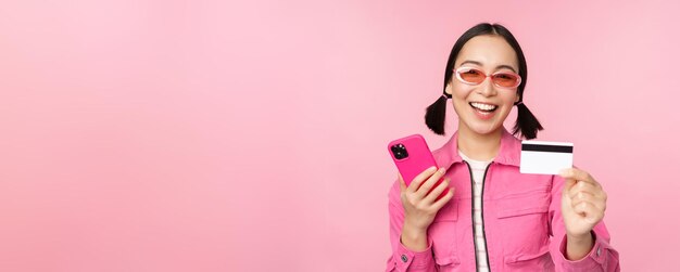 Linda garota asiática moderna rindo e sorrindo com cartão de crédito para celular compras on-line pagando com smartphone em pé sobre fundo rosa