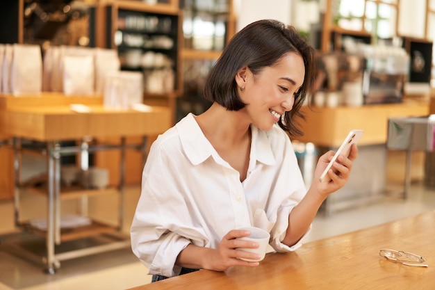 Linda garota asiática sorrindo e lendo no smartphone bebendo café sentado no café olhando para mim