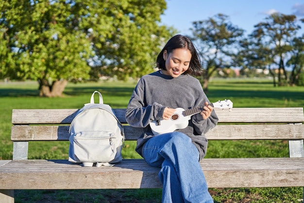 Linda garota asiática toca ukulele ao ar livre senta-se no parque no banco com instrumento musical