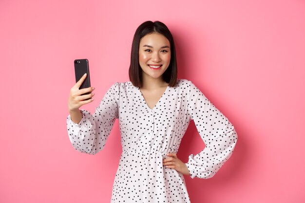 Linda garota asiática usando aplicativo de filtros de fotos e tomando selfie no celular, posando com um vestido fofo contra um fundo rosa.