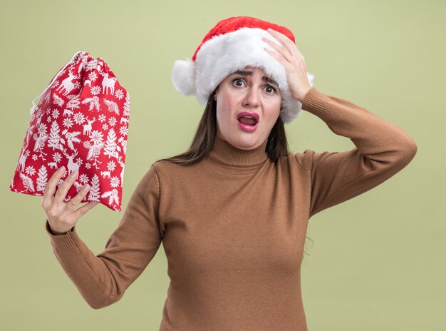 Linda garota assustada com chapéu de natal segurando uma sacola de natal e colocando a mão na cabeça isolada sobre fundo verde oliva