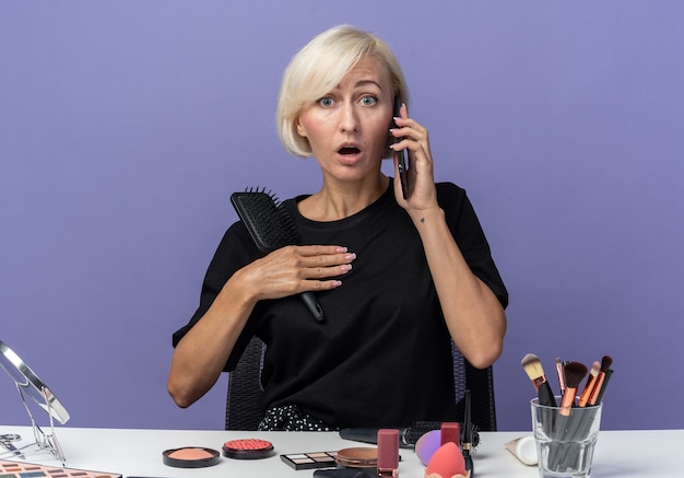 Linda garota assustada se senta à mesa com ferramentas de maquiagem falando no telefone segurando o pente isolado no fundo azul