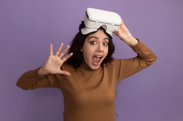 Linda garota assustada usando e segurando um fone de ouvido vr isolado na parede roxa