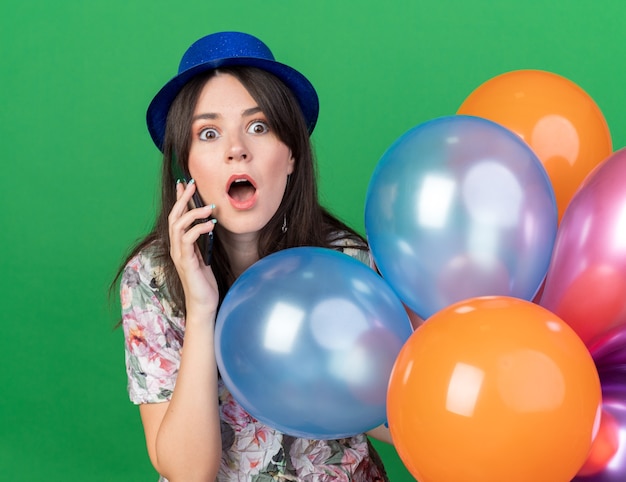 Linda garota assustada usando um chapéu de festa segurando balões falando ao telefone