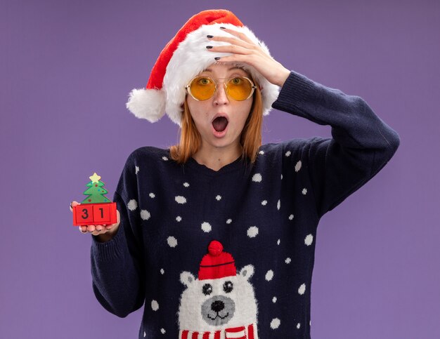 Foto grátis linda garota assustada vestindo um suéter de natal e um chapéu com óculos segurando um brinquedo de natal colocando a mão na testa isolada no fundo roxo