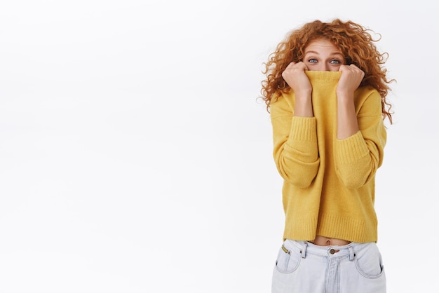 Linda garota boba se protege do inverno puxando o suéter amarelo aconchegante e quente no rosto ri e sorrindo com os olhos brincando no humor despreocupado brincalhão em pé fundo branco escondido