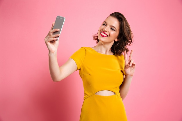 Linda garota bonita com maquiagem brilhante, mostrando o gesto de paz enquanto estiver a tomar selfie no telemóvel