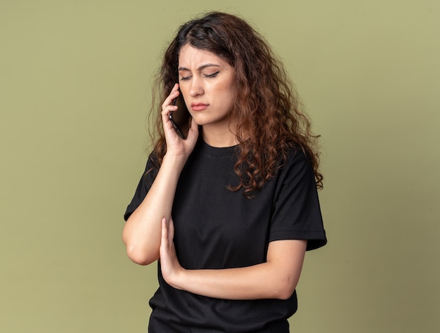 Linda garota carrancuda falando no telefone, olhando para baixo, isolada na parede verde oliva com espaço de cópia