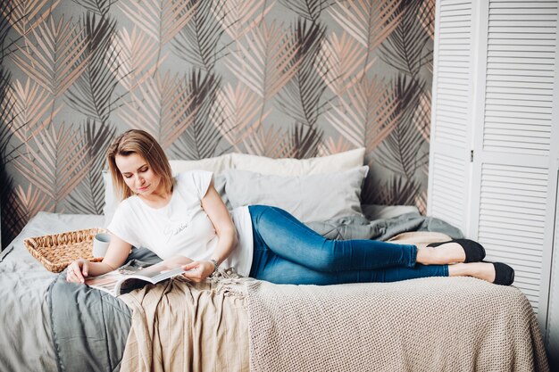 Linda garota caucasiana com cabelo claro, camiseta branca e jeans está no grande quarto bem iluminado e lê uma revista