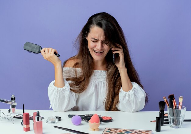 Linda garota chorando e sentada à mesa com ferramentas de maquiagem segurando um pente de cabelo falando no telefone olhando para baixo isolado na parede roxa