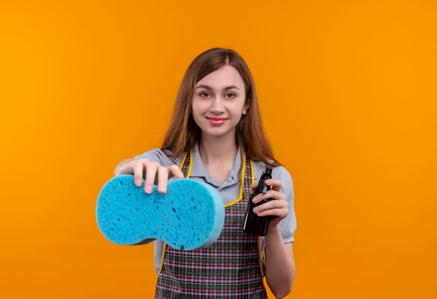 Linda garota com avental segurando spray de limpeza e esponja olhando para a câmera sorrindo, pronta para limpar