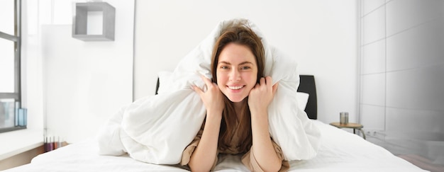 Linda garota com cabelo bagunçado, deitada na cama coberta com lençóis brancos, edredom, sorrindo e rindo coquete