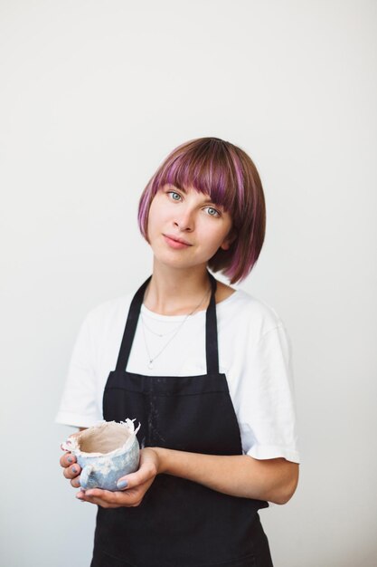 Linda garota com cabelo colorido no avental preto e camiseta branca segurando uma tigela artesanal sonhadoramente olhando na câmera no estúdio de cerâmica isolado